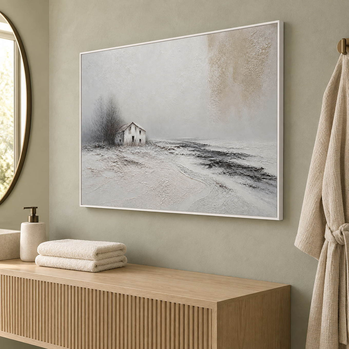 Winter landscape painting with a small house and dark trees beyond a snowy field in a white frame beside a bathroom vanity.