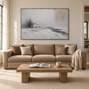 Winter landscape painting with a small house and dark trees beyond a snowy field in a brown wood frame in a living room.