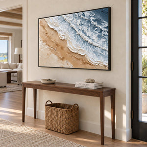 Kissing the Shore in a black frame above a wood entryway console with a woven basket beneath.