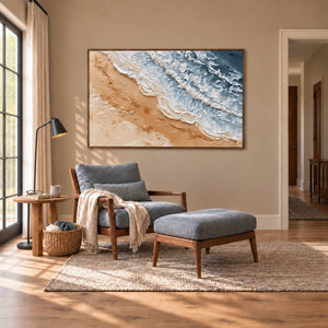 Kissing the Shore in a wood frame above a blue-gray armchair and matching footstool beside a window.