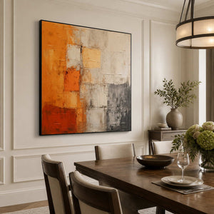 Black framed orange abstract painting beside a wooden dining table and flowers.