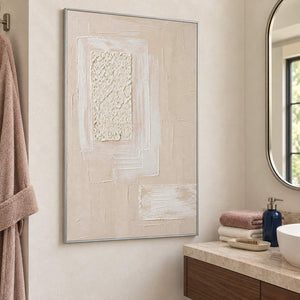 Ivory and blush relief painting with layered rectangles and a rough central panel in a silver frame beside a bathroom vanity.
