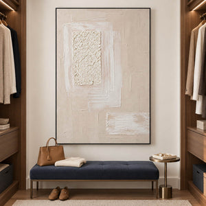 Ivory and blush relief painting with layered rectangles and a rough central panel in a black frame in a dressing room seating area.