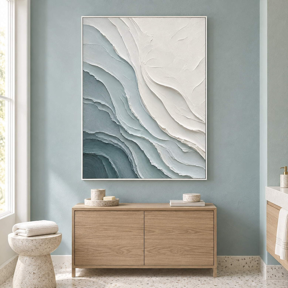 Layered blue-gray and chalk-white coastal relief painting in a white frame beside a bathroom vanity.