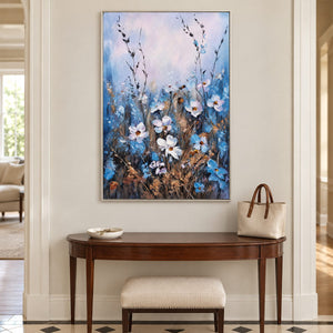 In an entryway above a console or bench, an artwork with a silver-colored frame shows white and blue wildflowers on slender stems beneath a broad pale upper area.