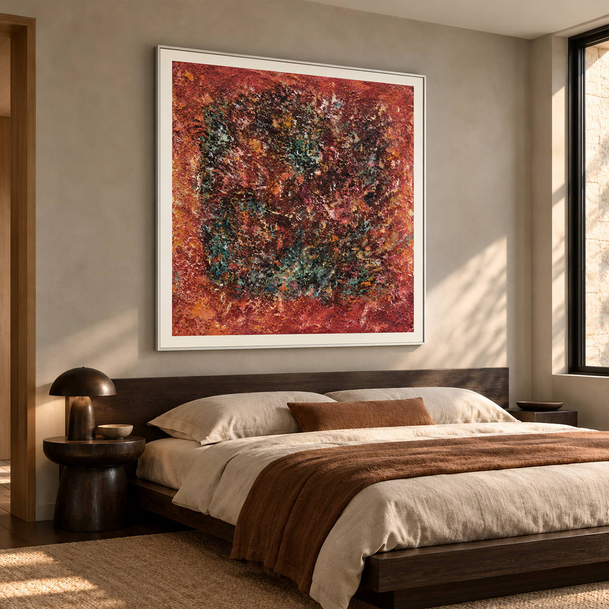 In a bedroom near the bed, an artwork with a white frame shows a dark textured ring surrounded by rust red and orange with scattered turquoise flecks.