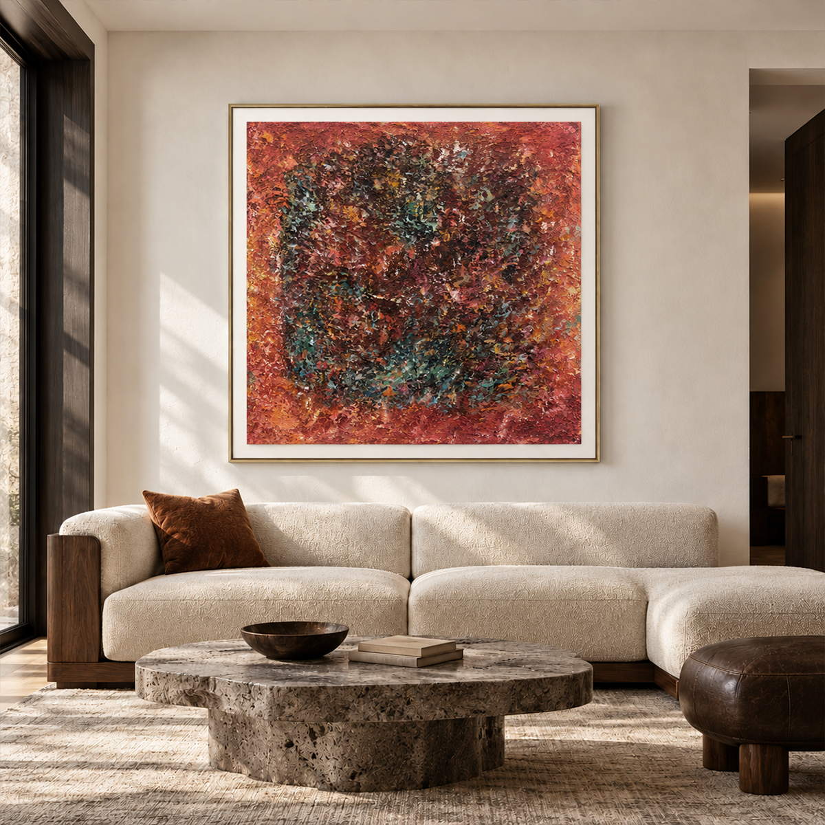 In a living room above seating, an artwork with a gold-colored frame shows a dark textured ring surrounded by rust red and orange with scattered turquoise flecks.