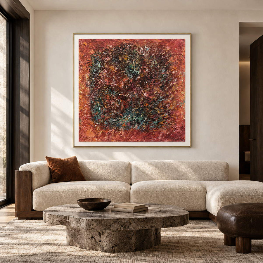 In a living room above seating, an artwork with a gold-colored frame shows a dark textured ring surrounded by rust red and orange with scattered turquoise flecks.