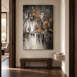 Dark gray and brown flaking forms above horizontal strata and pale vertical streaks in an artwork with no outer frame, displayed in a hallway above a bench.