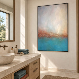 Turquoise coastal painting with a warm shoreline and a luminous pale sky in a black frame beside a bathroom vanity.