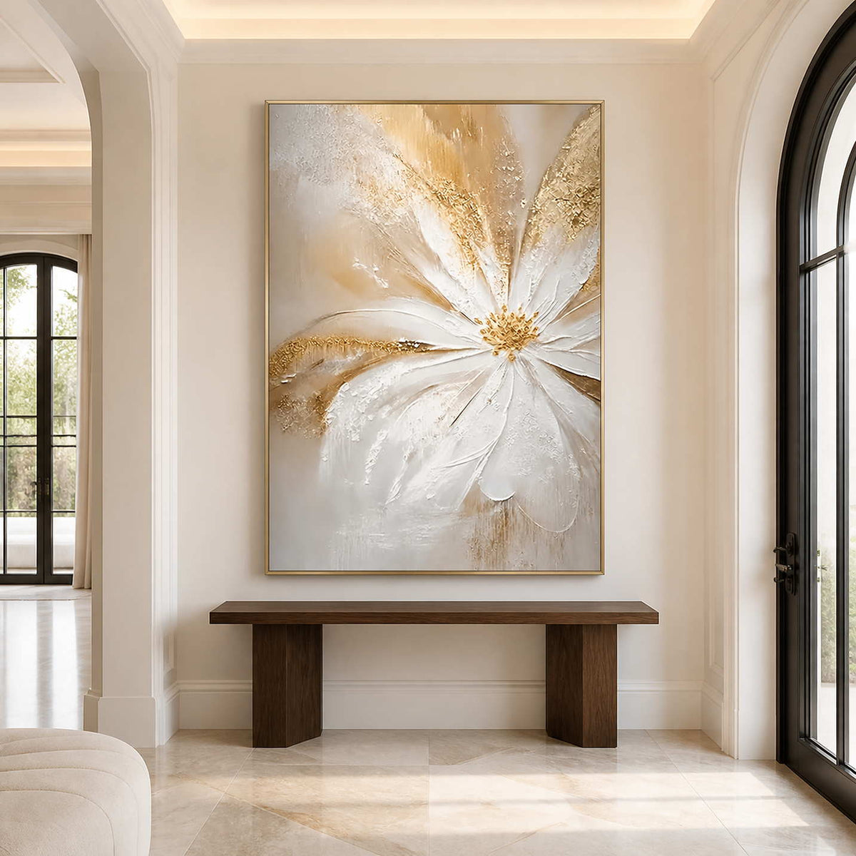 Gold-framed vertical white flower painting above a wood console in a bright entryway with arched windows.