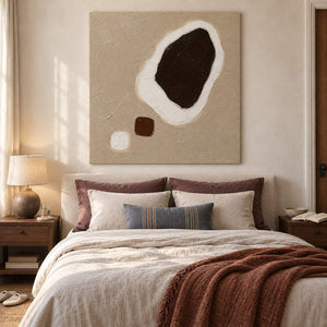 Taupe abstract painting with a large dark brown shape edged in white and two small forms without an outer frame in a bedroom.