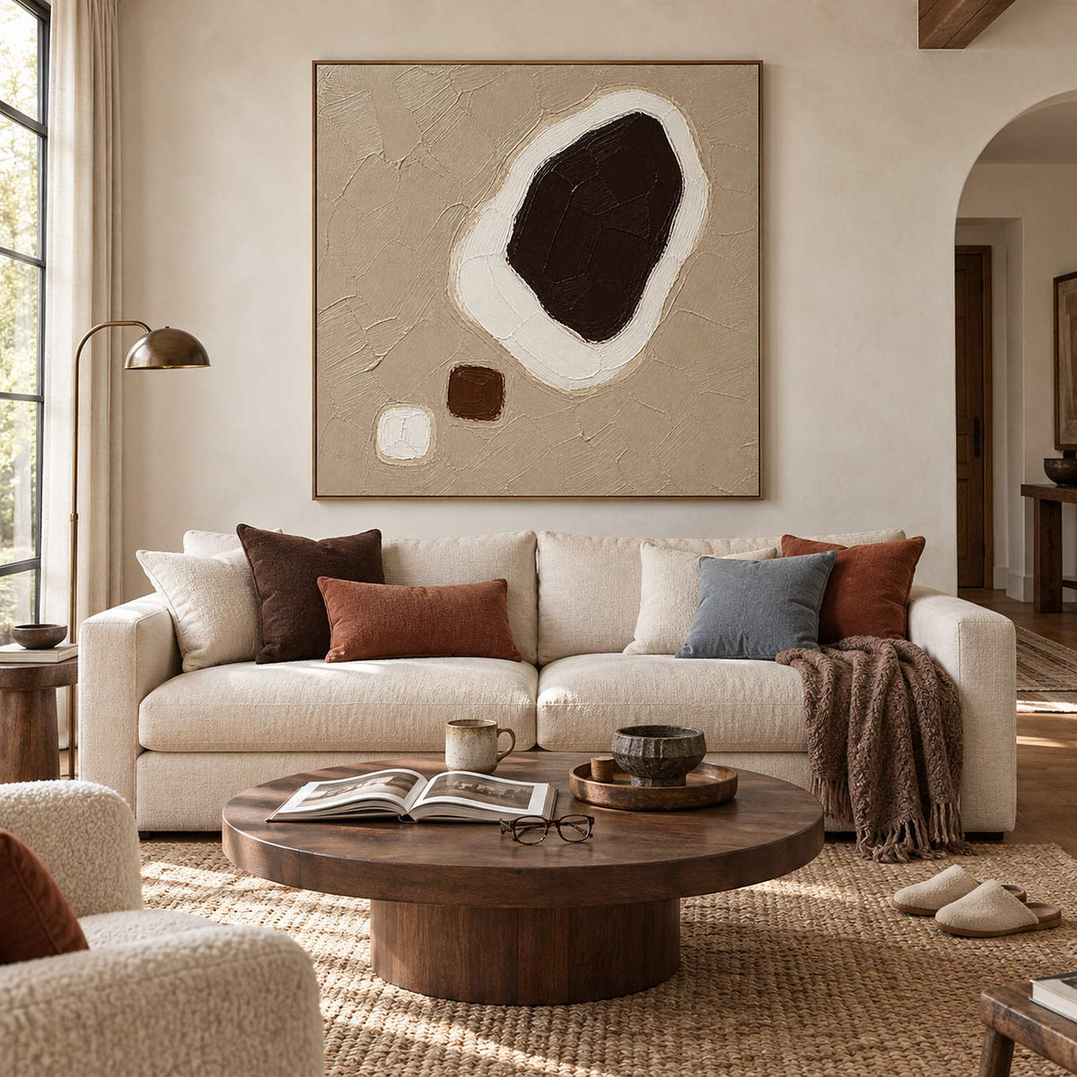 Taupe abstract painting with a large dark brown shape edged in white and two small forms in a brown wood frame in a living room.