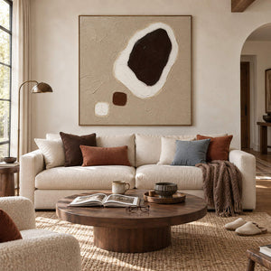 Taupe abstract painting with a large dark brown shape edged in white and two small forms in a brown wood frame in a living room.