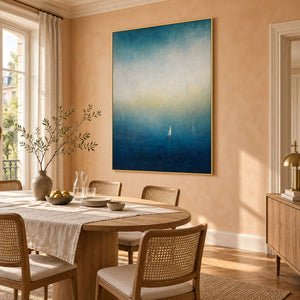 Atmospheric coastal painting with a white sail drifting across deep teal water, displayed in a dining room in a gold frame.