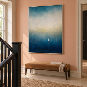 Atmospheric coastal painting with a white sail drifting across deep teal water, displayed beside a staircase in a silver frame.