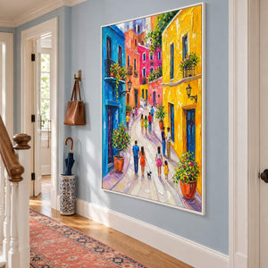 Colorful street painting with people, bright facades, and flowered balconies in a white frame in a hallway.