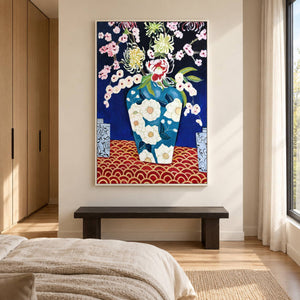 In a bedroom above a long bench near the bed, an artwork with a white frame shows flowers in a blue floral-patterned vase above a red surface with repeated gold-colored arcs.
