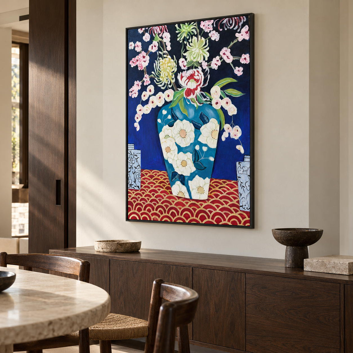 Artwork showing flowers in a blue floral-patterned vase above a red surface with repeated gold-colored arcs in a dining area beside the table, with a black frame.