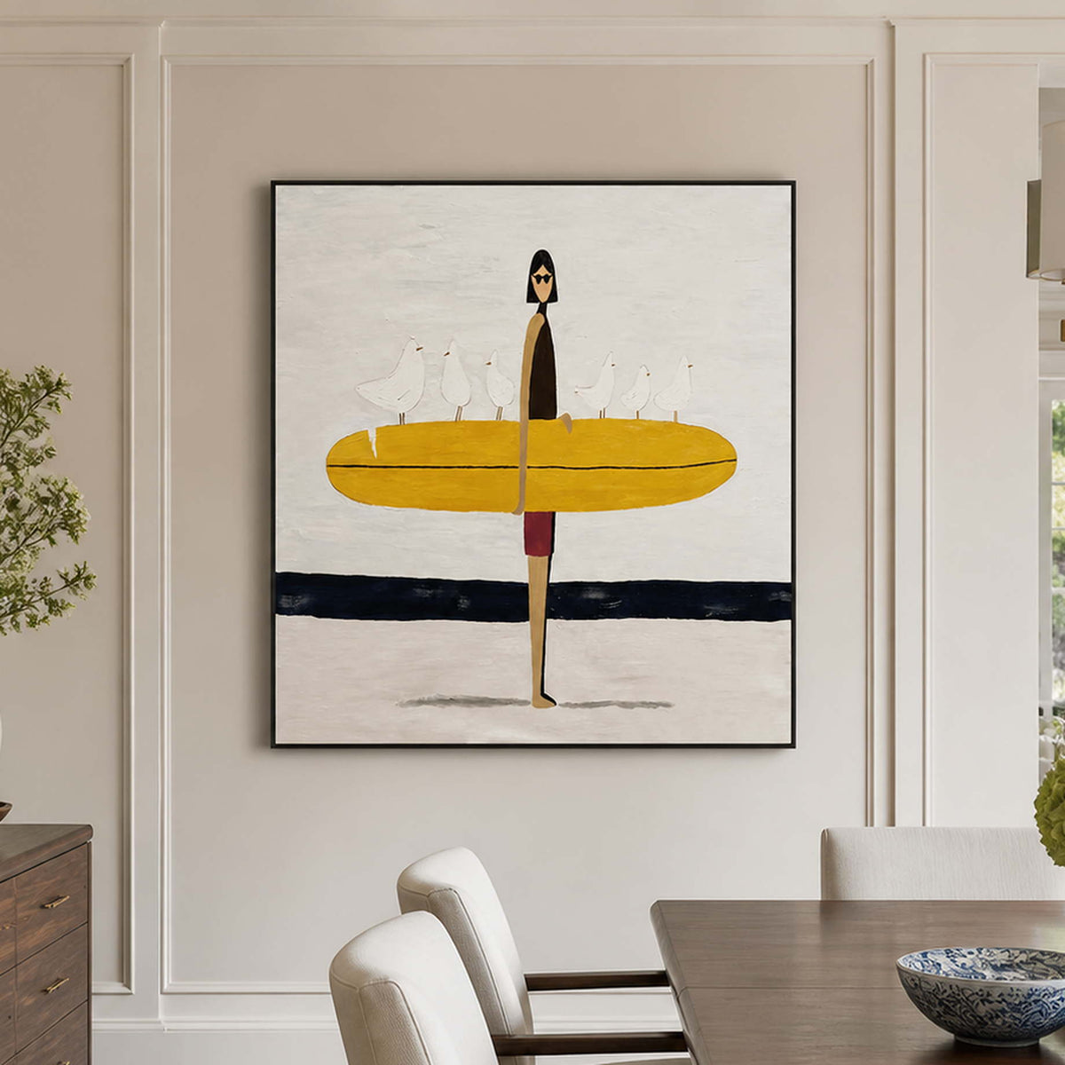 Black framed surfer artwork beside a wooden dining table with cream upholstered chairs.