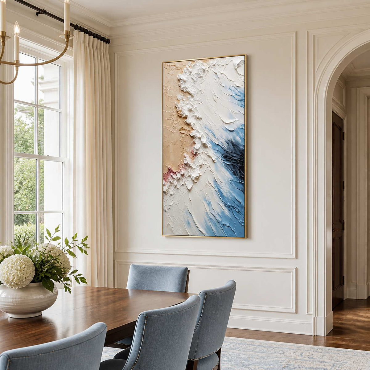 Gold-framed coastal abstract wall art above blue dining chairs beside a wooden table and window.