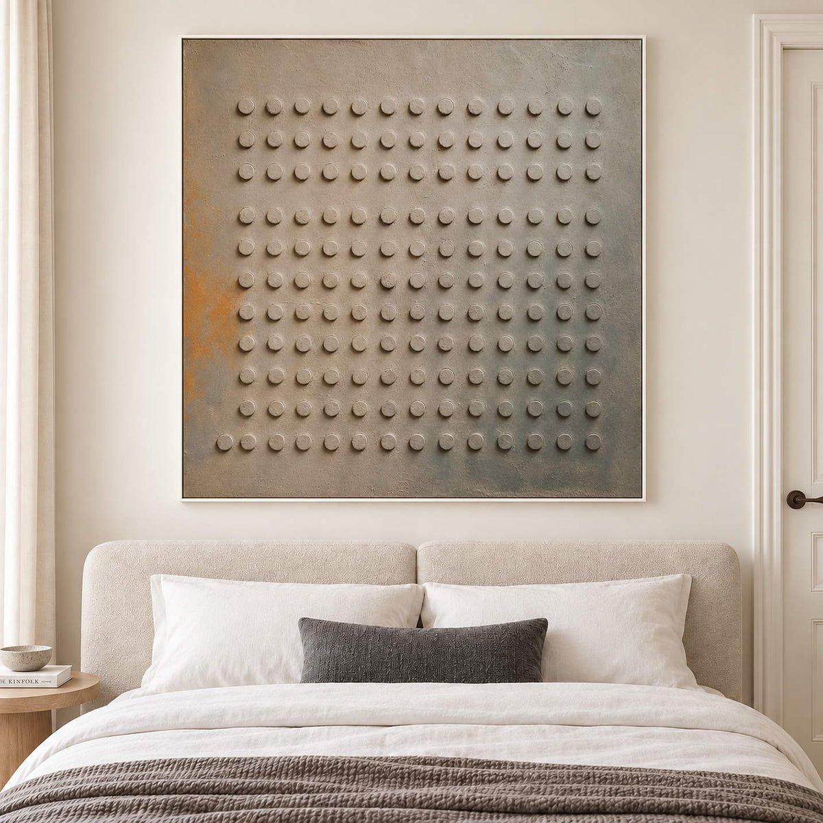 A regular grid of raised round forms on a gray earth-toned ground in a white frame, displayed above a bed.