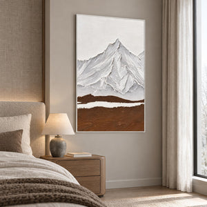 Artwork showing white and gray ridged mountains above a broad brown foreground and a narrow white band in a bedroom near the bed, with a white frame.