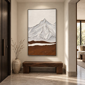 White and gray ridged mountains above a broad brown foreground and a narrow white band in an artwork with a black frame, displayed in an entryway above a console or bench.