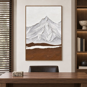 White and gray ridged mountains above a broad brown foreground and a narrow white band in an artwork with a brown wood-look frame, displayed in a study with a desk.