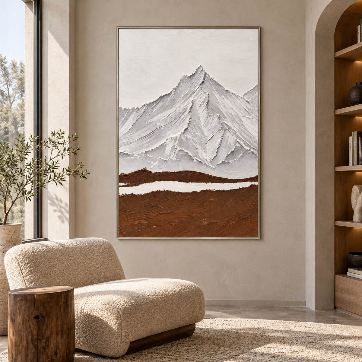 Beside a reading chair, an artwork with a silver-colored frame shows white and gray ridged mountains above a broad brown foreground and a narrow white band.