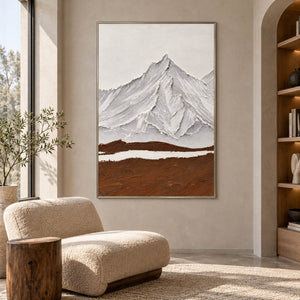 Beside a reading chair, an artwork with a silver-colored frame shows white and gray ridged mountains above a broad brown foreground and a narrow white band.