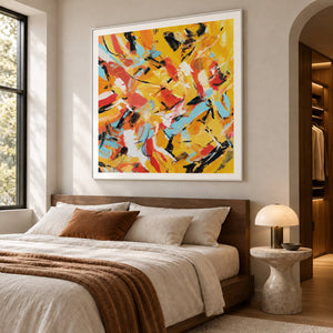 Interlocking yellow, orange, light blue, white and black abstract strokes in an artwork with a white frame, displayed in a bedroom near the bed.