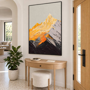 Black-framed mountain artwork beside an entry door, above a curved console table with a leafy plant to the left.
