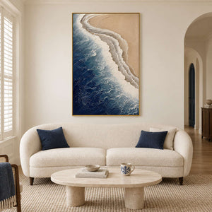 White surf curling along a sandy shore beside dark blue water in a gold frame, displayed above a sofa in a living room.