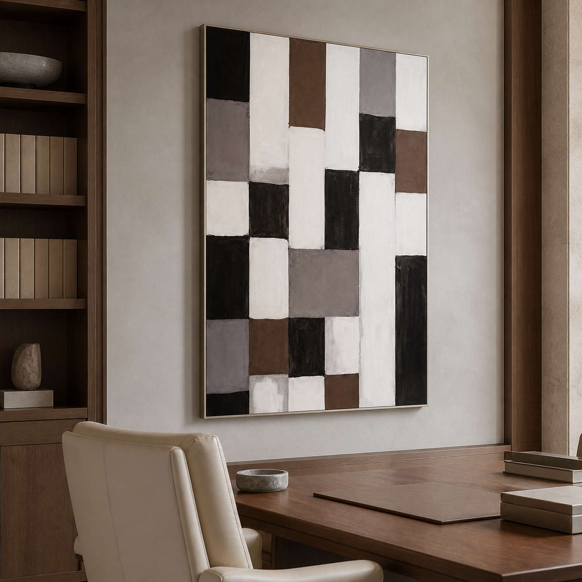 Silver framed geometric abstract artwork above a wooden desk beside bookshelves and a cream chair.