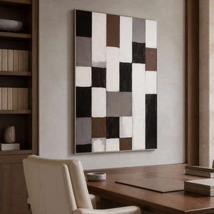 Silver framed geometric abstract artwork above a wooden desk beside bookshelves and a cream chair.