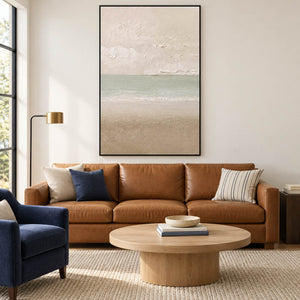 Hand-painted coastal canvas in warm ivory, sea-glass green, and sand, displayed in a living room in a black frame.