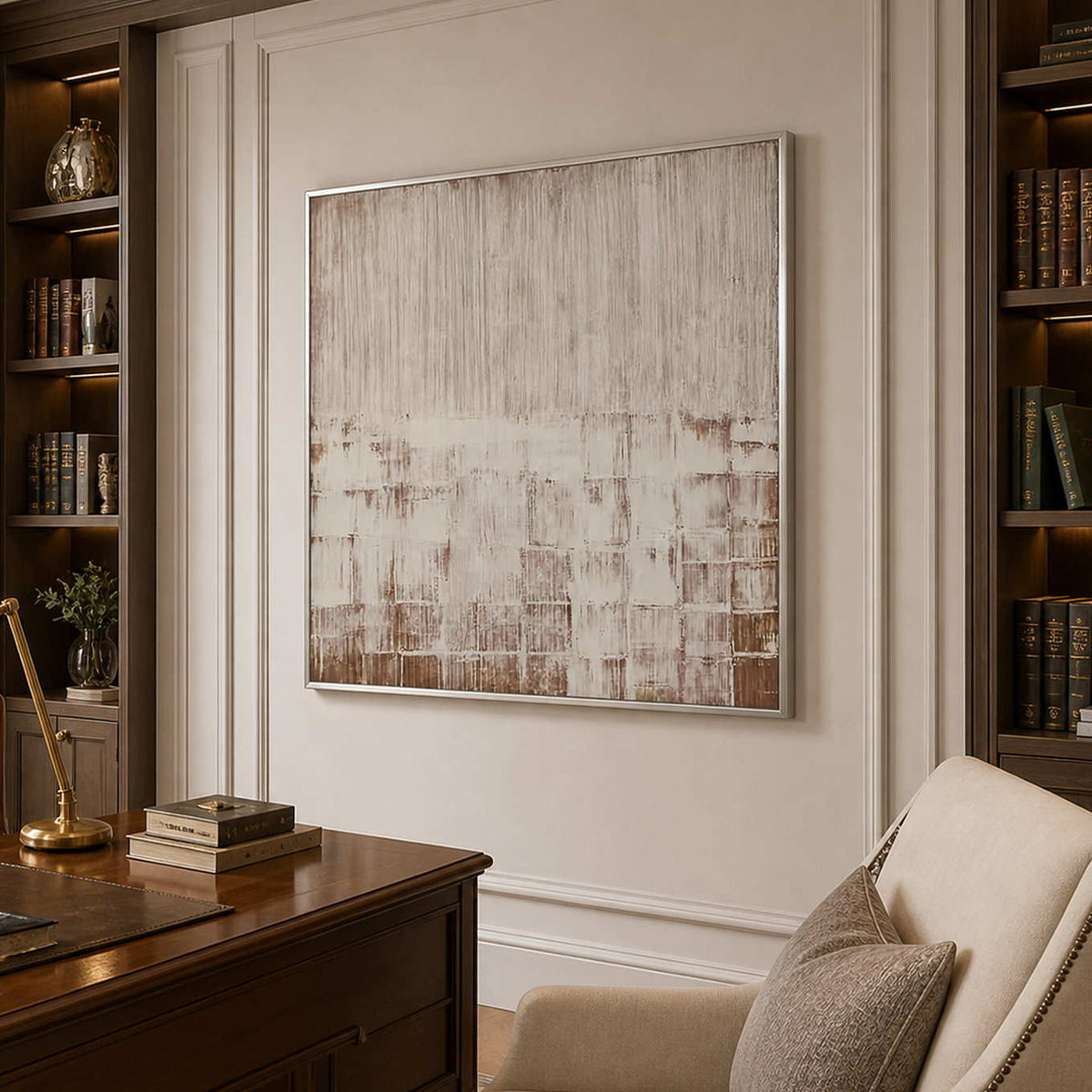 Silver framed cream and brown abstract artwork beside a wooden desk and upholstered chair.