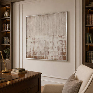 Silver framed cream and brown abstract artwork beside a wooden desk and upholstered chair.