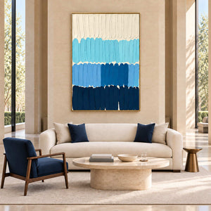 The Sea's Rhythm in a gold frame above a cream sofa and round stone table in a tall-windowed lobby.