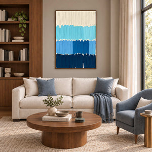 The Sea's Rhythm in a wood frame above a cream sofa with blue cushions and a round wood coffee table.