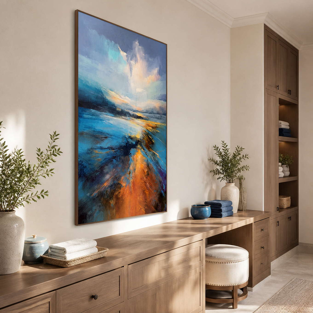 Wood framed blue landscape above bathroom cabinetry with folded towels and a padded stool.