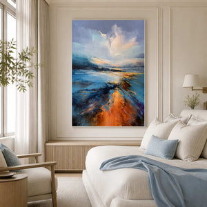 White framed blue landscape beside a cream bed with blue cushions and a blanket.