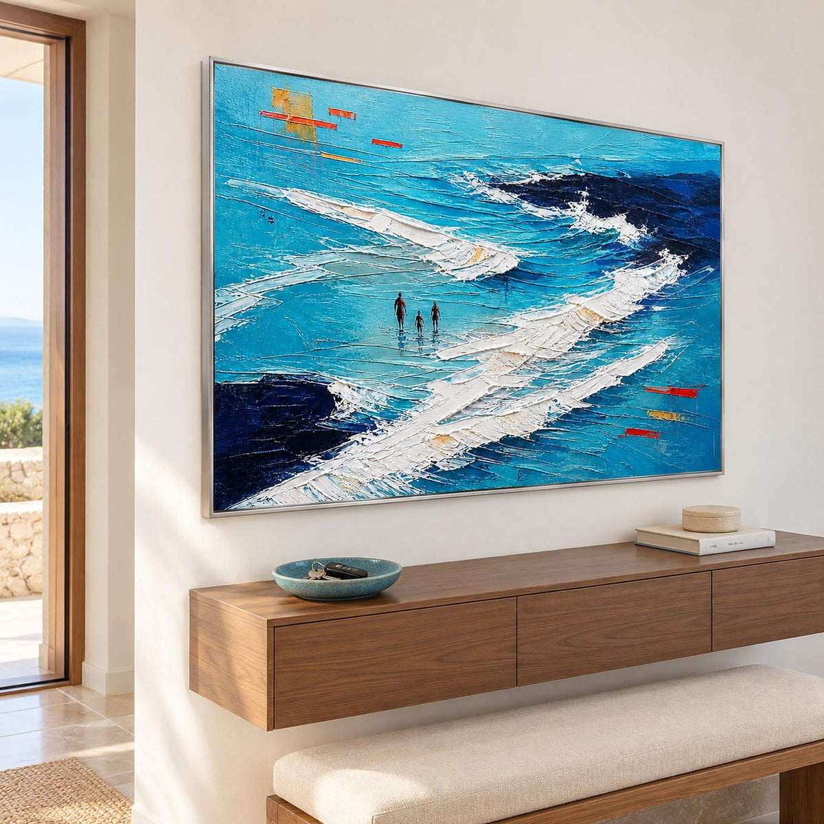Turquoise coastal painting with three figures among white breaking waves in a silver frame in an entryway.