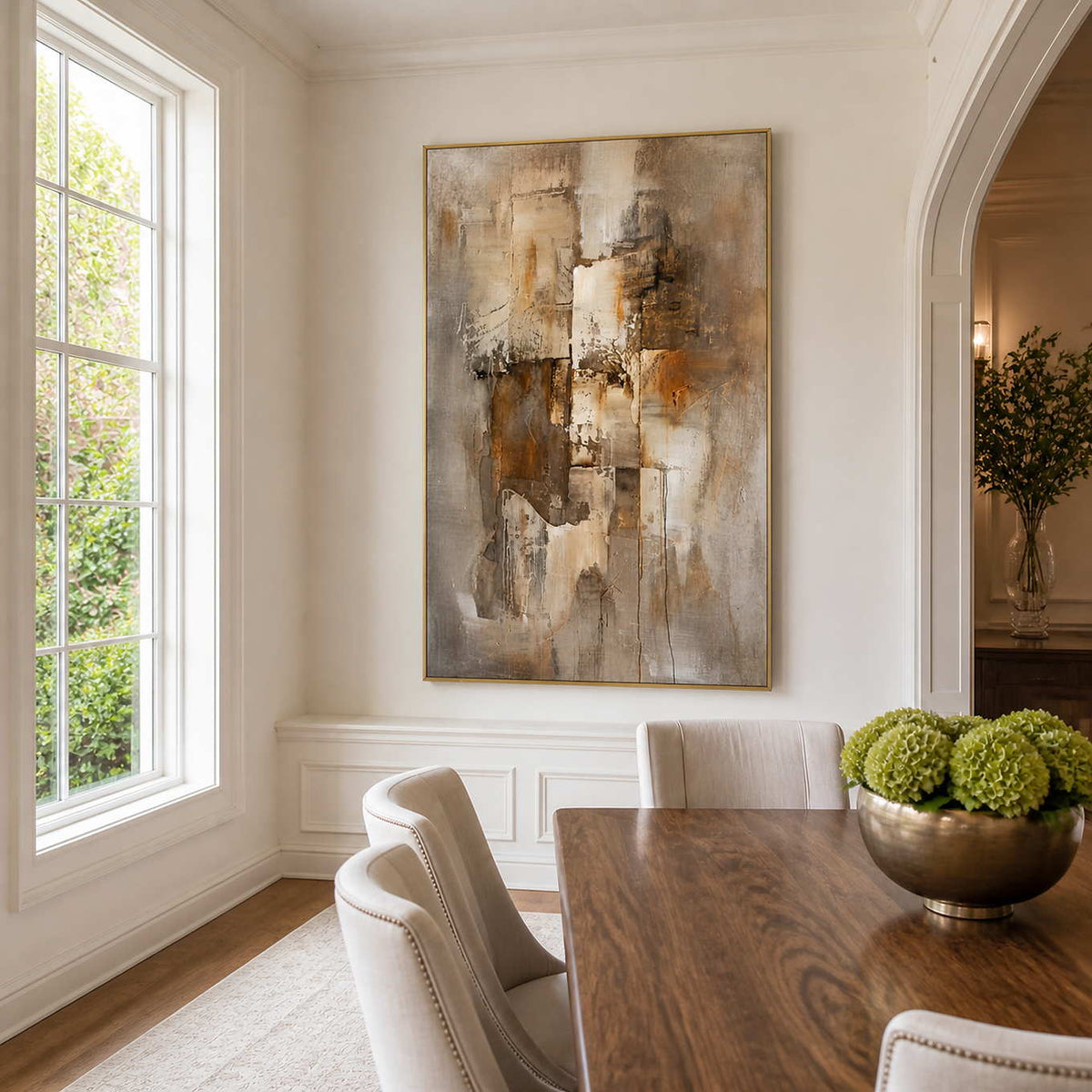 Gold-framed brown and taupe artwork beside a wooden dining table, cream chairs, and bright window.