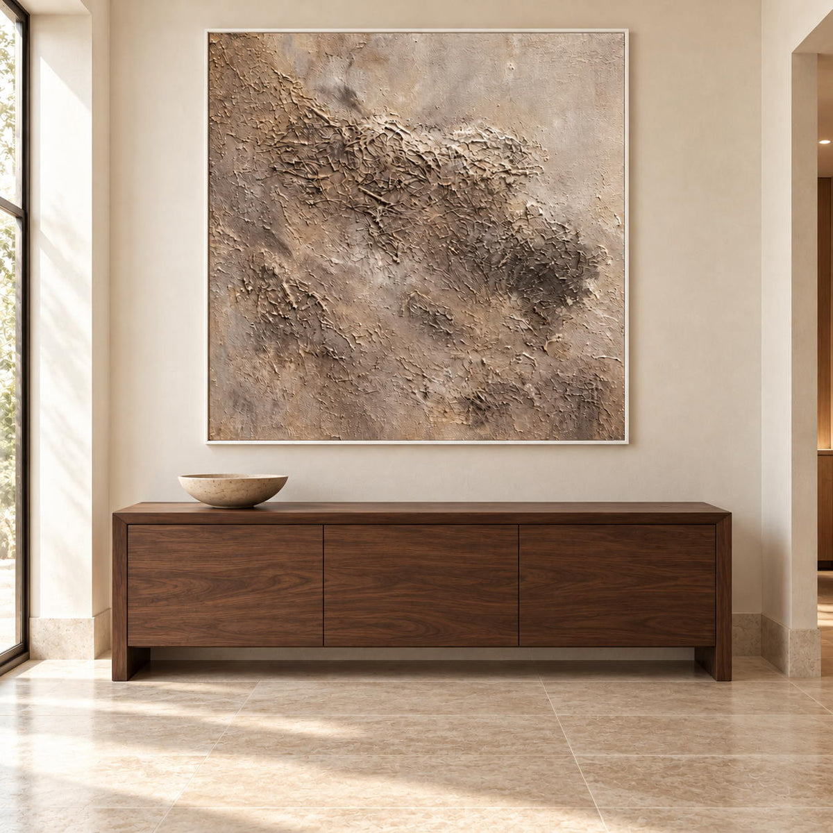 Above a low wooden sideboard, an artwork with a white frame shows irregular beige and taupe relief-like textures on a pale square ground.