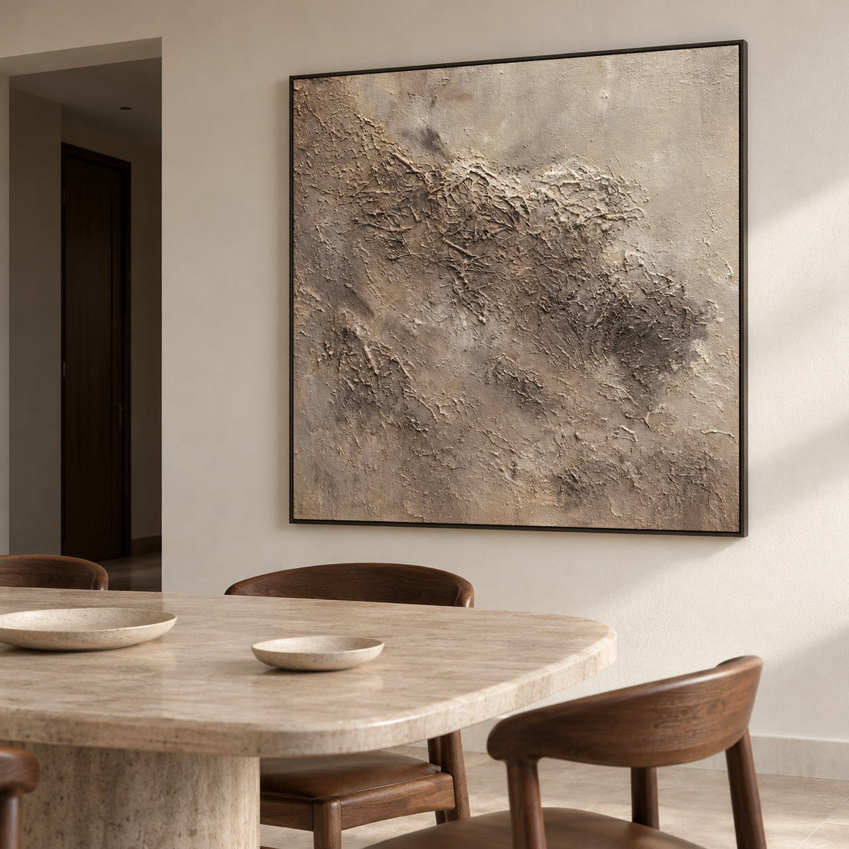 Artwork showing irregular beige and taupe relief-like textures on a pale square ground in a dining area beside the table, with a black frame.
