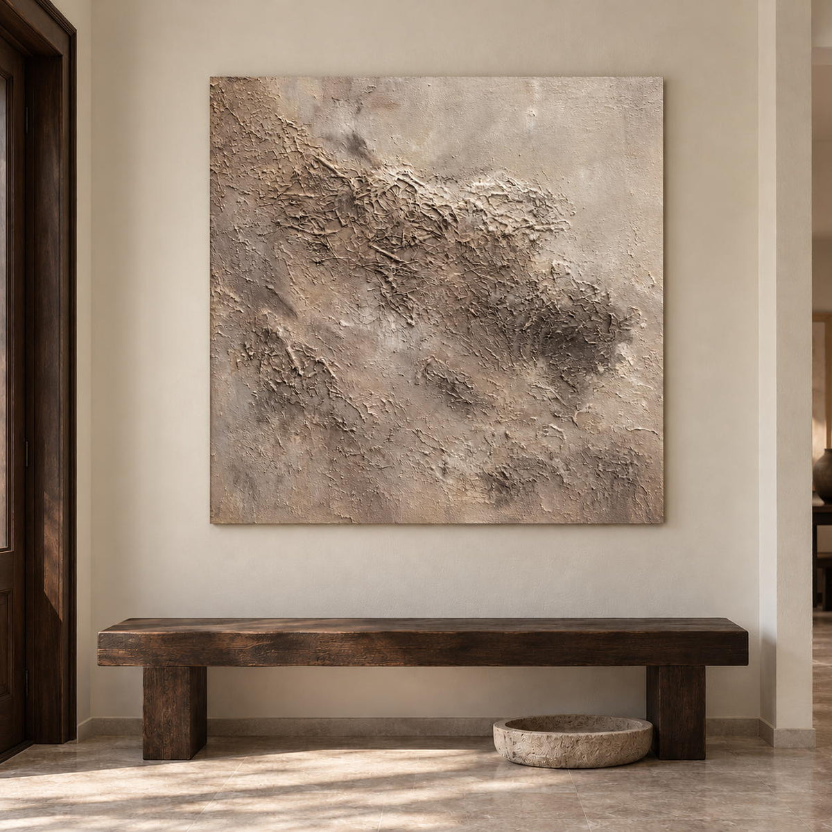 Artwork showing irregular beige and taupe relief-like textures on a pale square ground in an entryway above a console or bench, with no outer frame.