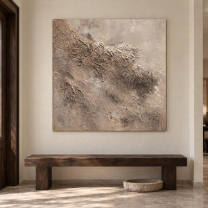 Artwork showing irregular beige and taupe relief-like textures on a pale square ground in an entryway above a console or bench, with no outer frame.