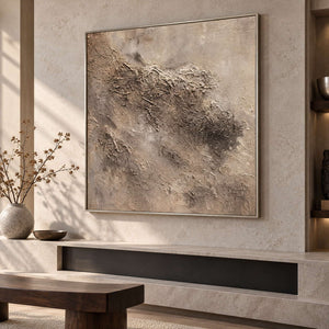 Irregular beige and taupe relief-like textures on a pale square ground in an artwork with a silver-colored frame, displayed above a fireplace.